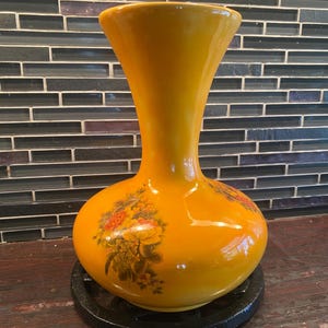 Può includere: Un vaso in ceramica giallo senape lucido con un bordo largo e svasato e una base arrotondata. Il corpo del vaso presenta una stampa floreale decorativa nei toni dell'arancione, del giallo e del verde, raffigurante fiori in piena fioritura.