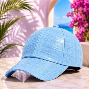 May include: A light blue baseball cap featuring a textured crocodile skin pattern across the entire surface. The cap has a structured crown, a curved brim, and a button at the top centre, showcasing a consistent embossed reptile scale design.