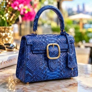 May include: A structured handbag featuring a vibrant blue snakeskin texture. The bag includes a single top handle, a front flap closure secured by a prominent gold-toned rectangular buckle, and a matching leather tag hanging from the side.