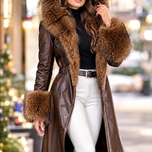 May include: A long brown leather coat featuring a large hood and cuffs trimmed with thick brown fur. The coat is worn open over a black mock neck top and white trousers, cinched at the waist with a black belt featuring a gold-toned buckle.
