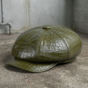 May include: An olive green newsboy cap featuring a textured crocodile skin pattern across the entire surface. The hat has a rounded, multi-paneled crown construction with a small button at the top center and a short, stiff brim extending forward.