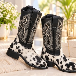 Peut inclure: Une paire de bottes mi-mollet avec un motif imprimé vache noir et blanc sur la partie inférieure et le pied. La tige est noire avec des surpiqûres décoratives crème formant un motif de feuilles tourbillonnantes et des tirettes en haut.