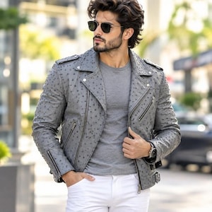 Puede incluir: Una chaqueta de moto de cuero gris texturizado con solapa de muesca, cremalleras de metal plateado y charreteras, usada sobre una camiseta gris lisa y pantalones blancos. La chaqueta presenta un patrón de puntos distintivo en toda la superficie.