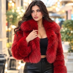 May include: A vibrant red faux fur jacket with a thick, plush collar and long sleeves, worn over a simple black crop top and black leather-style trousers. The jacket features a dense, textured pile and an open front design with a cropped length.