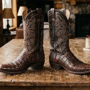 Op de afbeelding: Een paar donkerbruine leren cowboylaarzen met een textuur van krokodillenleer op de voet en een hoge schacht met ingewikkelde decoratieve stiksels. De laarzen hebben een puntige neus, een hak en treklussen aan de bovenranden.