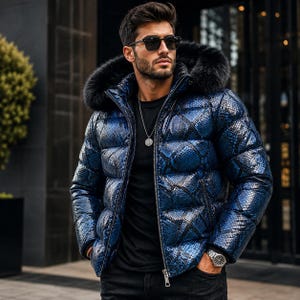 K&ouml;nnte beinhalten: Eine Daunenjacke mit gl&auml;nzendem blau-schwarzem Schlangenmuster, einer Kapuze mit schwarzem Fellbesatz, einem Frontrei&szlig;verschluss und langen &Auml;rmeln. Die Jacke wird &uuml;ber einem schlichten schwarzen T-Shirt getragen, kombiniert mit einer schwarzen Hose.