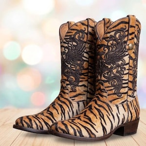 Op de afbeelding: Een paar halfhoge cowboylaarzen met een tijgerstreepmotief in tinten oranje, zwart en beige. De laarzen zijn versierd met gedetailleerd donkerbruin borduurwerk op de schacht en hebben een kleine metalen stud aan de zijkant.
