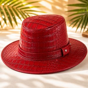 May include: A vibrant red hat featuring a glossy, embossed crocodile skin texture throughout. The hat has a flat top, a wide brim, and a matching red band around the base of the crown, secured with a small, polished silver-toned metal stud.