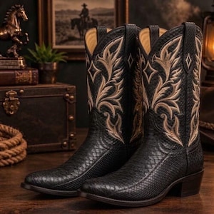 Puede incluir: Un par de botas vaqueras negras con un patrón texturizado similar a la piel de serpiente en el pie y la caña. La parte superior de la caña está decorada con un intrincado bordado de color tostado en forma de hoja, con un diseño de diamante.