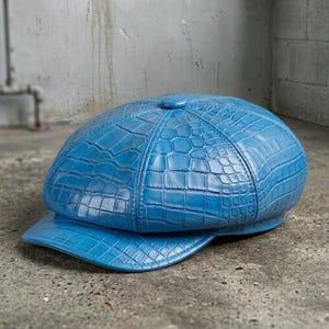 May include: A bright blue newsboy cap featuring a textured crocodile skin pattern across the entire surface. The hat has a rounded, multi-paneled crown construction with a small button at the top center and a short, curved brim at the front.