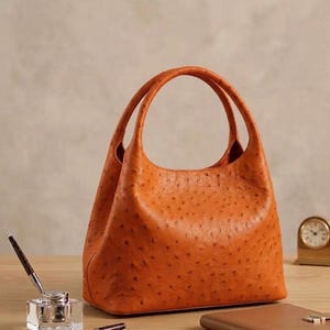 May include: A burnt orange handbag featuring a distinct textured surface with small, evenly spaced indentations. The bag has a rounded top opening and two integrated handles. The design is structured with a wide base and tapers toward the top.