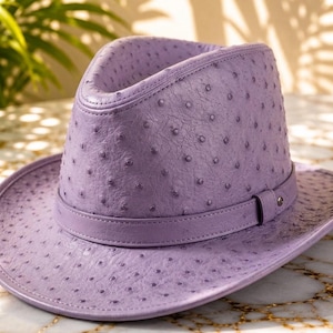 May include: A lavender fedora hat featuring a textured ostrich skin pattern with raised quill bumps. The hat includes a matching lavender band around the base of the crown and a structured brim that curves slightly upward at the sides.