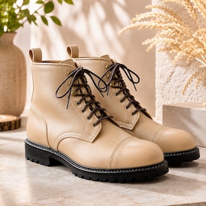 May include: A pair of beige ankle boots featuring a smooth finish, a rounded toe cap, and dark brown laces threaded through brass-toned eyelets. The boots have a sturdy black lug sole and a pull tab at the back of the collar for easy access.