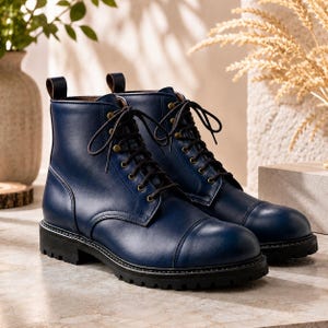 May include: A pair of navy blue leather boots featuring a cap toe design, dark brown laces, and brass-toned eyelets. The boots have a sturdy black lug sole and a pull tab at the heel, providing a structured silhouette with visible stitching details.