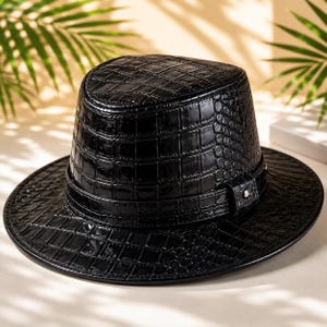 Peut inclure: Un chapeau fedora noir avec une finition brillante et texturée façon peau de crocodile. Le chapeau présente une bande noire assortie autour de la base de la calotte, agrémentée d'un petit détail en métal argenté centré sur le côté.