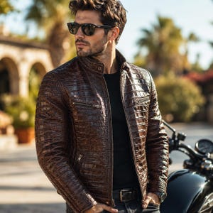 May include: A dark brown leather jacket with a textured crocodile skin pattern, featuring a stand-up collar, a full-length front zip, and two horizontal zipped chest pockets. The jacket is worn over a plain black shirt with dark trousers.