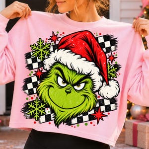 Könnte beinhalten: Ein hellrosa Sweatshirt mit Rundhalsausschnitt und einem Grafikdruck einer grünen Figur mit roter Weihnachtsmütze. Das Design zeigt einen schwarz-weiß karierten Hintergrund, hellgrüne Schneeflocken und rote Sterne als Akzente.