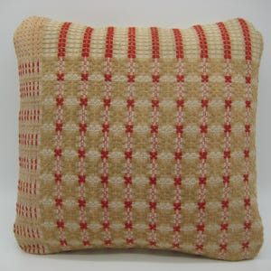 May include: A square throw pillow featuring a woven texture with a grid pattern in tan and beige. The design includes vertical stripes of bright red thread woven into the fabric, creating a repeating cross-stitch effect across the surface.