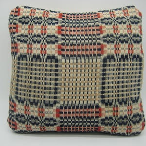 May include: A square throw pillow featuring a woven plaid pattern in shades of navy blue, cream, and burnt orange. The fabric displays a textured, grid-like design with repeating geometric blocks and vertical and horizontal interlaced threads.
