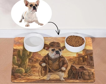 May include: A rectangular pet feeding mat featuring a French Bulldog dressed as a cowboy in a desert town setting, accompanied by two white ceramic bowls, one empty and one filled with brown dry kibble, with a circular inset showing the dog.