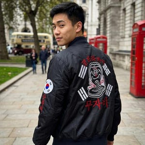 Op de afbeelding: Een zwart bomberjack met een grote afbeelding op de achterkant van een cobra omringd door elementen van de Zuid-Koreaanse vlag en Koreaanse tekst. De mouw heeft een ronde patch van de Zuid-Koreaanse vlag met rode Koreaanse tekst eronder.