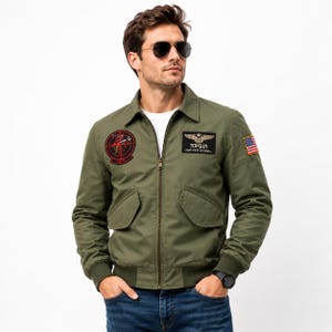 May include: An olive green bomber jacket featuring a front zipper, ribbed cuffs, and two flap pockets. The chest displays a circular red and black patch and a rectangular black patch with the text TOP GUN CAPT-PETE MITCHELL. A small American flag patch is on the sleeve.