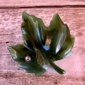 May include: A green leaf-shaped dish with a glossy finish. The dish has a small, round, silver-colored embellishment. The leaf has detailed veining and a stem. The dish is likely made of glass or a similar material.