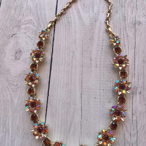 May include: A gold-tone metal necklace featuring a series of flower-shaped links set with amber-colored rectangular stones and iridescent, multi-colored faceted crystals. The necklace includes a chain extension with a hook clasp closure.