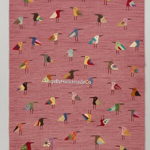 May include: A rectangular rug with a dusty rose background featuring a repeating pattern of small, stylized birds in various colors and geometric designs. The text RugsByHandmadeCo is printed in white across the center of the rug surface.