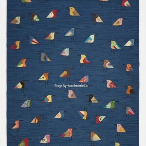 Puede incluir: Una alfombra rectangular con fondo azul profundo que presenta un patrón repetitivo de pequeños pájaros estilizados en varios colores, incluyendo amarillo, rosa, verde, naranja y rojo. El texto RugsByHandmadeCo está impreso en blanco en el centro.