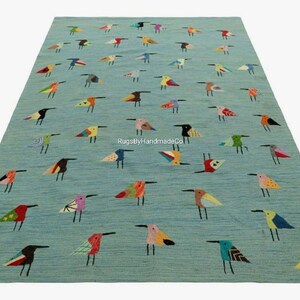 Puede incluir: Una alfombra rectangular con fondo azul claro que presenta un patrón repetitivo de pequeños pájaros estilizados en varios colores, incluyendo rojo, amarillo, negro y verde. El texto RugsByHandmadeCo está impreso en blanco en el centro.