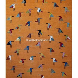 Puede incluir: Una alfombra rectangular de color naranja con un patrón repetitivo de pequeños pájaros estilizados en varios colores y diseños geométricos. El texto RugsByHandmadeCo está impreso en blanco en el centro de la alfombra entre las aves.