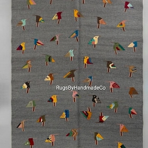 May include: A rectangular grey rug featuring a repeating pattern of small, stylized birds in various colours including yellow, blue, red, green, and brown. The text "RugsByHandmadeCo" is printed in white across the centre of the rug surface.