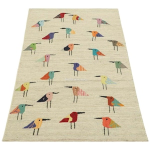 Puede incluir: Una alfombra rectangular con fondo beige claro que presenta un patrón repetitivo de aves estilizadas en varios colores, incluyendo rojo, azul, verde, naranja y amarillo. Cada ave está representada con líneas simples y formas geométricas.
