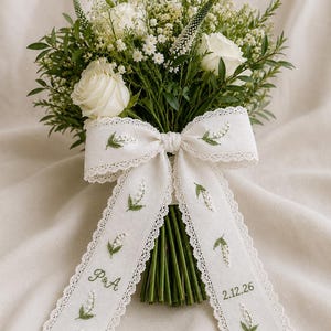 Op de afbeelding: Een boeket van witte rozen, kleine witte bloemen en groen blad, samengebonden met een wit stoffen lint. Het lint is voorzien van geborduurde lelietjes-van-dalen, de initialen P & A en de datum 2.12.26, afgewerkt met een fijne kanten rand.