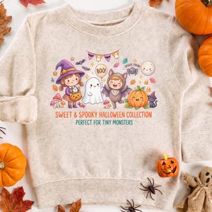 May include: A beige crewneck sweatshirt featuring a colorful Halloween graphic of a child in a witch costume, a ghost, and a child in a bat costume. The text reads SWEET & SPOOKY HALLOWEEN COLLECTION and PERFECT FOR TINY MONSTERS in orange and teal.