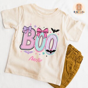 May include: A cream-colored short-sleeve t-shirt featuring the word BOO in striped pink, teal, and purple letters. The design includes pink and purple bows, small bats, and stars. The name Charlie is printed in pink script below the word BOO.
