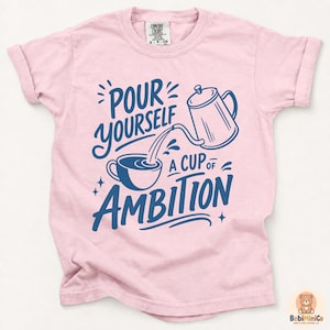 May include: A light pink short-sleeve t-shirt featuring a dark blue graphic print of a coffee pot pouring into a cup. The text reads, "POUR YOURSELF A CUP OF AMBITION." The shirt has a crew neckline and a Comfort Colors brand label at the inner collar.
