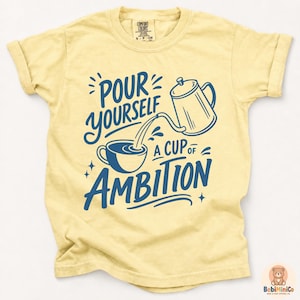 May include: A pale yellow short-sleeve t-shirt featuring a dark blue graphic print of a coffee pot pouring into a cup. The text reads, "POUR YOURSELF A CUP OF AMBITION." The shirt has a crew neckline and a Comfort Colors brand label at the inner collar.