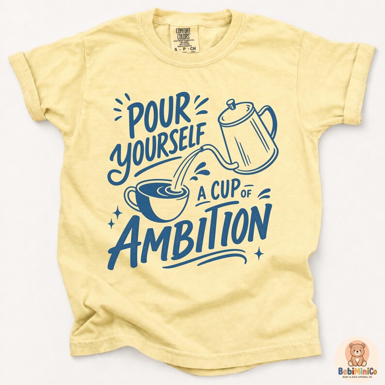 May include: A pale yellow short-sleeve t-shirt featuring a dark blue graphic print of a coffee pot pouring into a cup. The text reads, "POUR YOURSELF A CUP OF AMBITION." The shirt has a crew neckline and a Comfort Colors brand label at the inner collar.