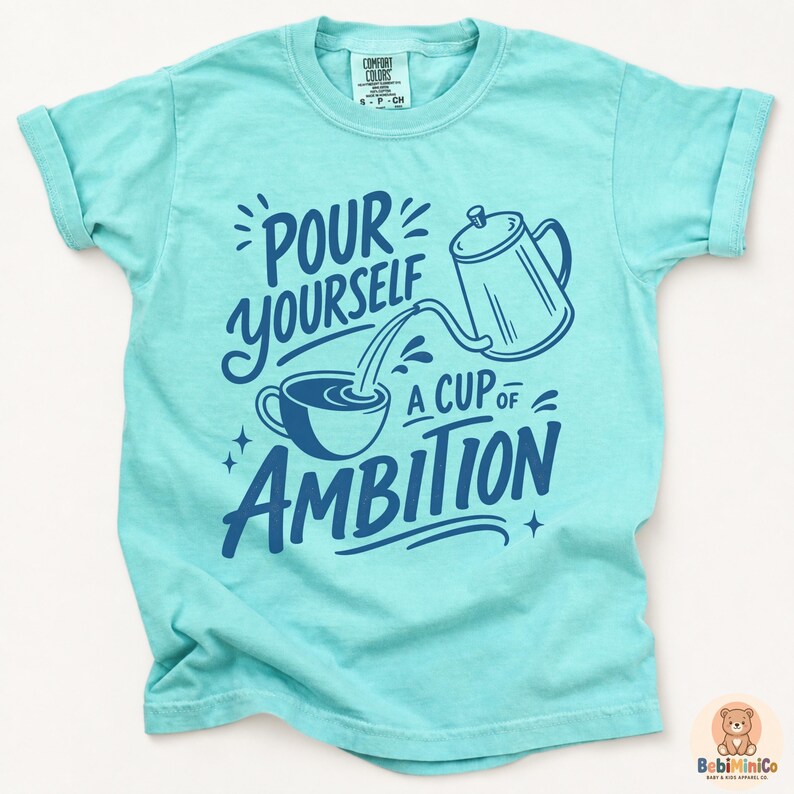 May include: A light turquoise short-sleeve t-shirt featuring a dark blue graphic of a coffee pot pouring into a cup with the text "POUR YOURSELF A CUP OF AMBITION" printed in a stylized font. The shirt has a crew neckline and a Comfort Colors label.