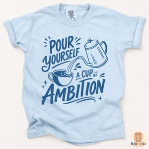 May include: A light blue short-sleeve t-shirt featuring a dark blue graphic of a coffee pot pouring into a cup with the text "POUR YOURSELF A CUP OF AMBITION" in a stylized font. The shirt has a crew neckline and a Comfort Colors brand label.