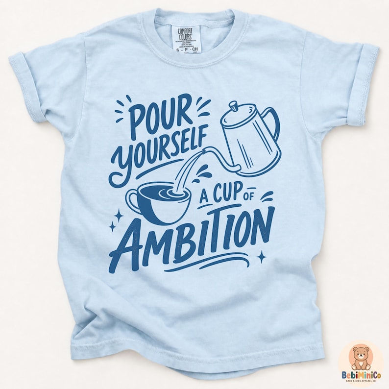 May include: A light blue short-sleeve t-shirt featuring a dark blue graphic of a coffee pot pouring into a cup with the text "POUR YOURSELF A CUP OF AMBITION" in a stylized font. The shirt has a crew neckline and a Comfort Colors brand label.