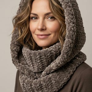 May include: A thick, plush, taupe-colored hooded cowl made of chunky, soft-textured yarn. The accessory features a deep, rounded hood that drapes around the neck and shoulders, providing a layered, cozy appearance with a consistent knit pattern.