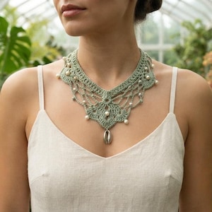 May include: A sage green crochet necklace featuring an intricate lace-like pattern, embellished with small white beads throughout the design, and finished with a single silver-toned cowrie shell pendant hanging from the center of the piece.
