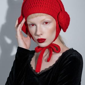 Puede incluir: Un gorro de ganchillo rojo brillante que presenta dos cuernos de diablo prominentes y erguidos en la coronilla y solapas circulares para las orejas en los lados. El gorro se asegura bajo la barbilla con dos lazos largos de ganchillo rojo.
