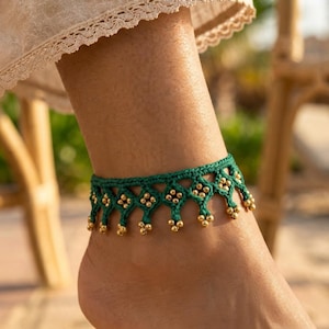 Puede incluir: Una tobillera de macramé verde oscuro con un patrón de diamantes tejidos, con pequeñas cuentas metálicas doradas integradas en los nudos y colgando del borde inferior, creando un efecto de flecos decorativos alrededor del tobillo.