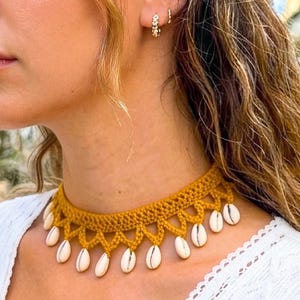 May include: A mustard yellow crocheted choker necklace featuring a scalloped edge design. The necklace is adorned with multiple small, white, oval-shaped cowrie shells hanging from the bottom points of the crocheted fabric pattern.