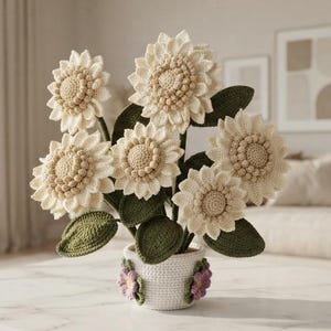 May include: A crochet flower arrangement featuring six cream-colored sunflowers with textured centers and dark green leaves. The flowers are contained in a white crocheted pot decorated with small purple and green floral accents at the base.