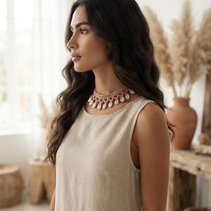 May include: A dusty pink crocheted choker necklace featuring a series of small, white, oval-shaped cowrie shells dangling from the bottom edge. The necklace is constructed with a delicate, intricate thread pattern and sits snugly around the neck.